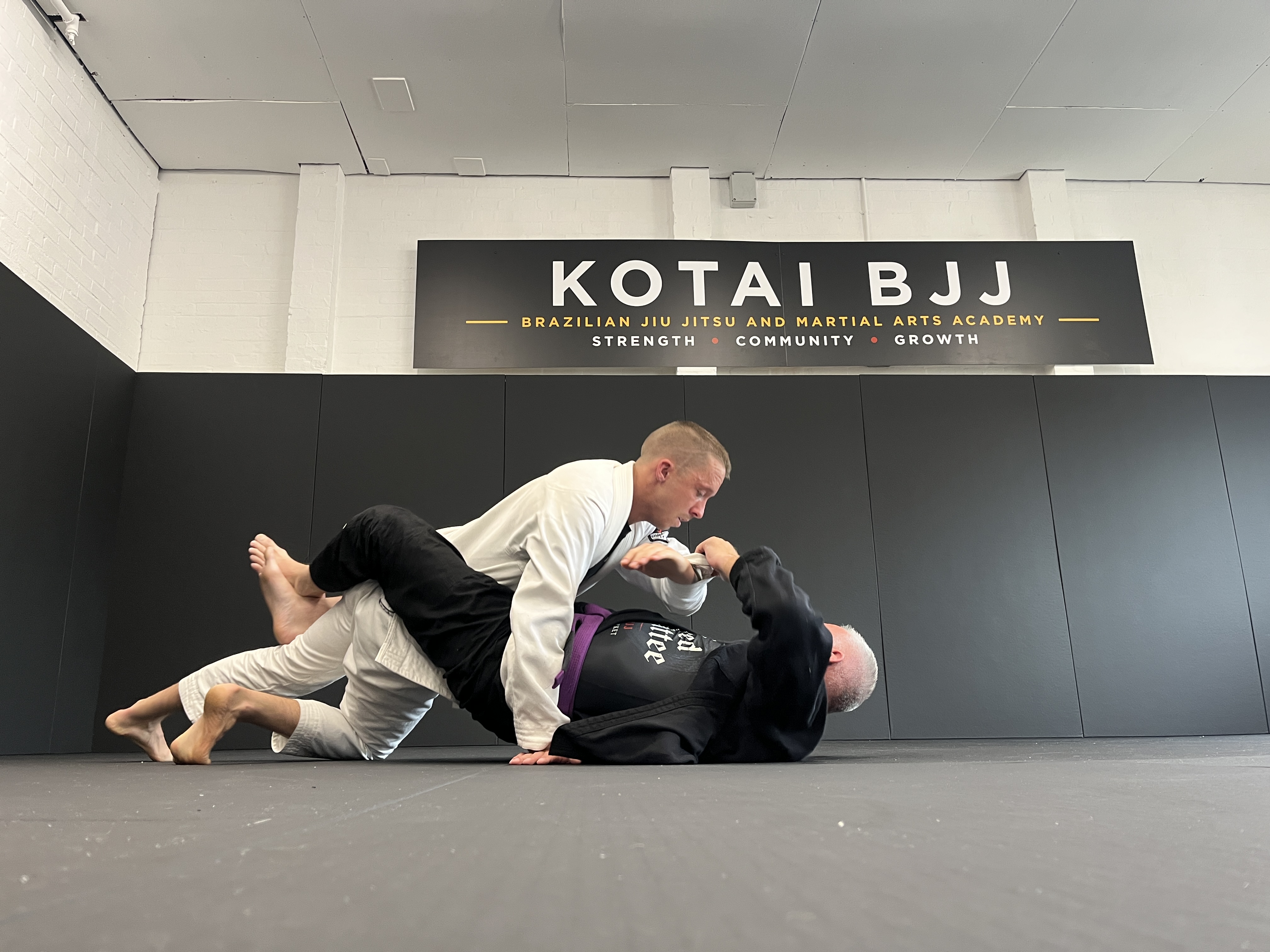 Two members sparring in the gi on the mats beneath the Kotai BJJ sign
